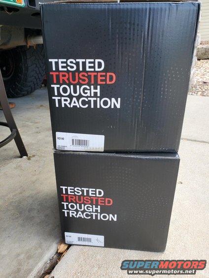 Two black boxes stacked, each saying 'TESTED TRUSTED TOUGH TRACTION'. Part of a vehicle tire and bumper visible to the left