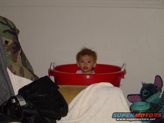 Baby in a red bucket with a stuffed toy and clothes nearby