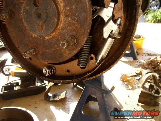Close-up of a rusty brake drum assembly with springs and brake cylinder. Some text is visible on the cylinder