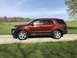 2016 Ford Explorer - This orange SUV is parked on a gravel road with a grassy field and trees in the background under a clear blue sky.