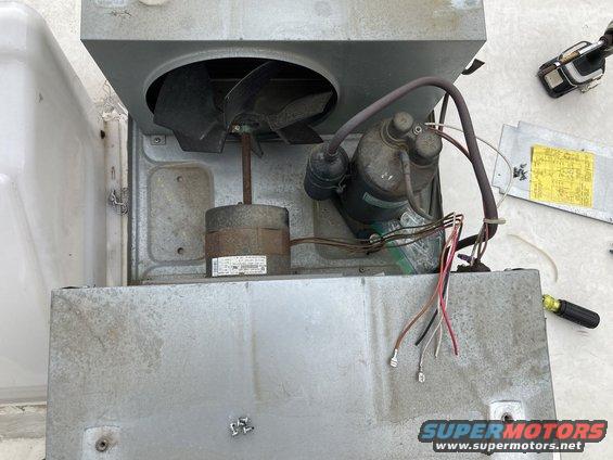 Air conditioner unit with fan, compressor, and exposed wiring. A yellow diagram is visible in the background