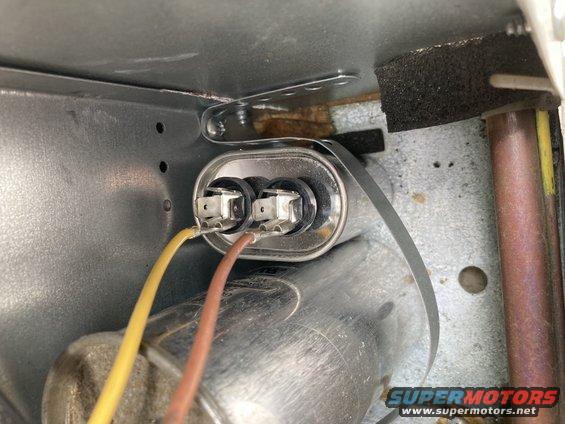 Capacitor with yellow and brown wires connected, secured by a metal strap inside a metal enclosure
