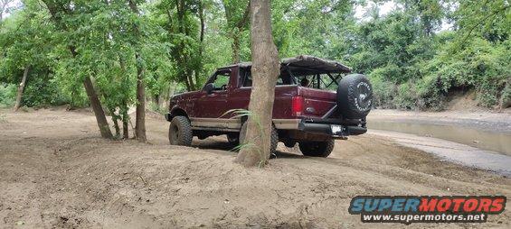 Maroon Ford Bronco with spare tire on back, driving on dirt path near trees and water