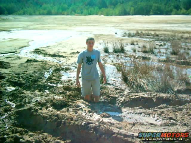 Person kneels in thick mud with a body of water and trees in the background