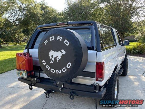 Rear view of a white Ford Bronco with a black spare tire cover showing the Bronco logo and a red gas can mounted on the back