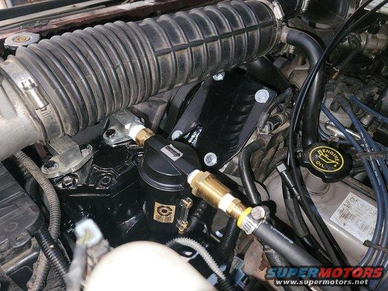 Engine bay with black air compressor, hoses, and air intake tube. Visible text includes "ENGINE OIL" and "SUPERMOTORS.NET"