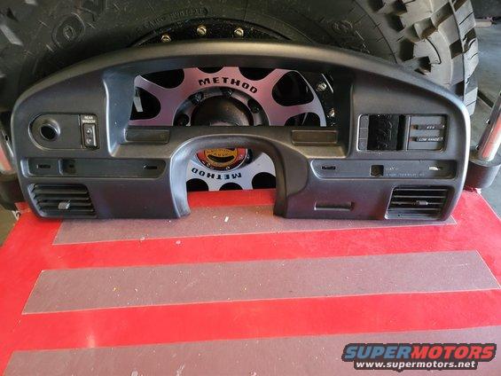 Dark gray dashboard instrument cluster with air vents, radio slot, and switches, behind a tire and Method wheel