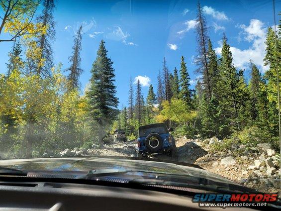 Blue SUV on rocky mountain road, rear view. Supermotors.net logo visible