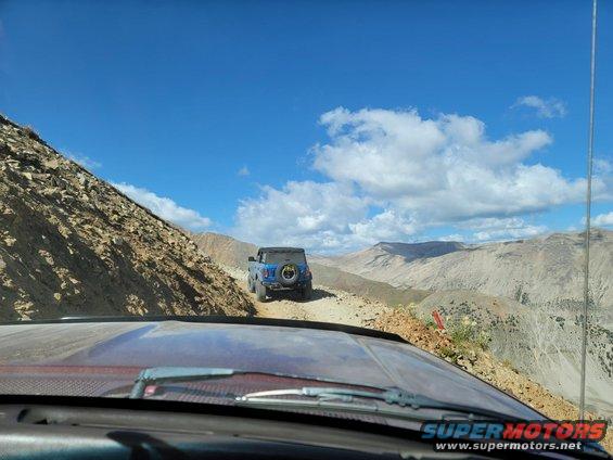 View from inside a vehicle looking at a blue SUV ahead on a dirt mountain road with rocky slopes and distant mountains under a blue sky with clouds