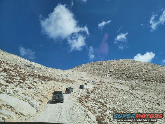 Multiple dark SUVs ascend a gravelly mountain path under a blue sky with scattered clouds