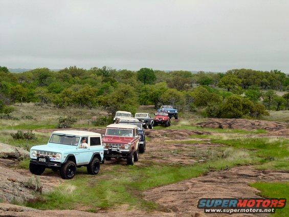 Light blue 1973 Ford Bronco with a white top, driving on a rocky, grassy terrain