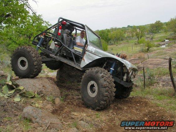 Silver 1973 Ford Bronco modified for off-roading, climbing a rock with large tires and a roll cage