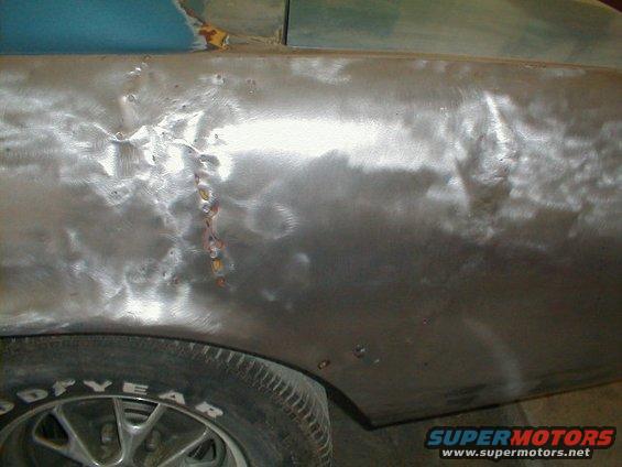 Car's rear quarter panel is bare metal, showing dents and weld repairs. A Goodyear tire is visible
