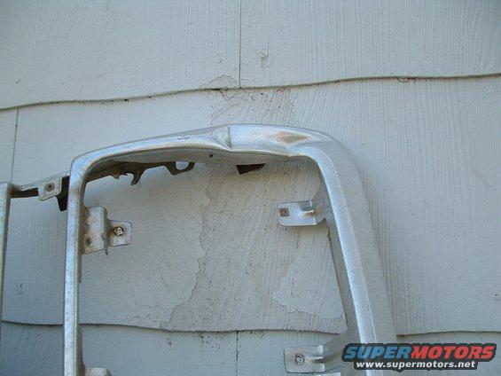 Chrome grille surround with dents and peeling paint, mounted against a white clapboard wall