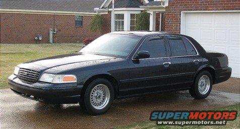 Black sedan parked in front of a brick house with a white garage door. The car has aftermarket wheels