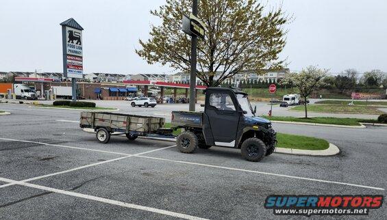 Dark gray UTV with a trailer attached, parked in a parking lot. The UTV has a cab and a cargo bed