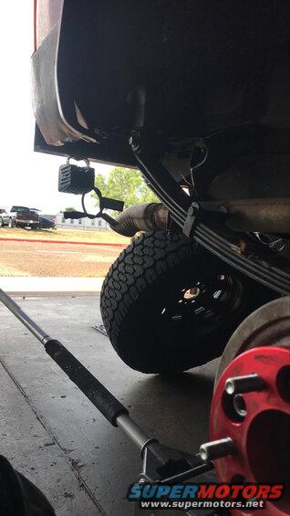 Close-up of a vehicle's rear axle, leaf springs, and tire. A red brake rotor is visible in the foreground