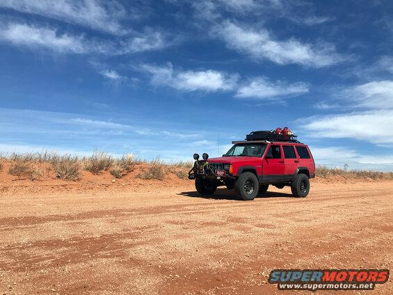 Red 1996 Jeep Cherokee with roof rack and off-road lights parked on a dirt road under a blue sky with scattered clouds
