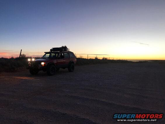 Red Jeep Cherokee with roof rack parked on dirt road at dusk