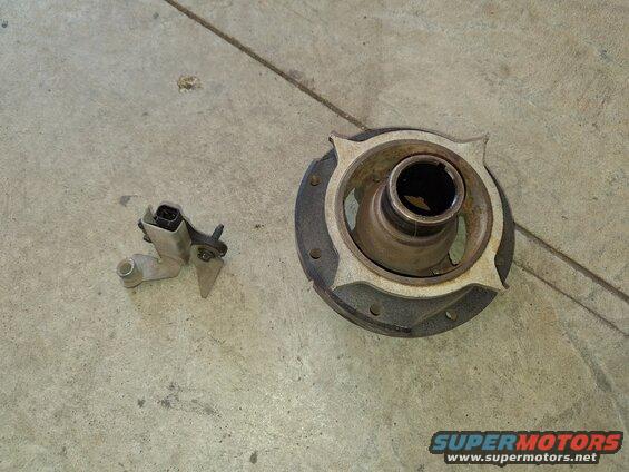 Two automotive parts on a concrete floor: a metal sensor with a black connector and a circular metal component with a central opening