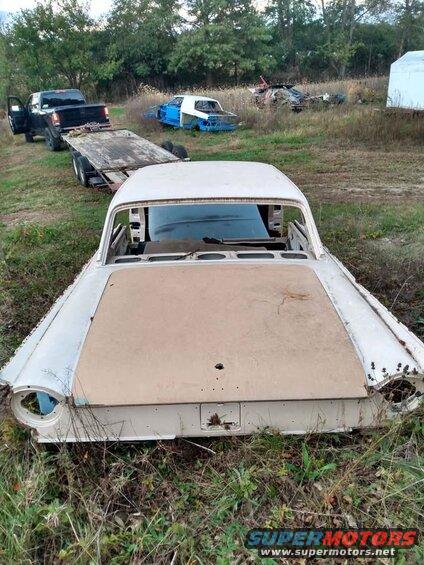 Rear view of a white 1960s car body with a trailer and pickup truck in the background