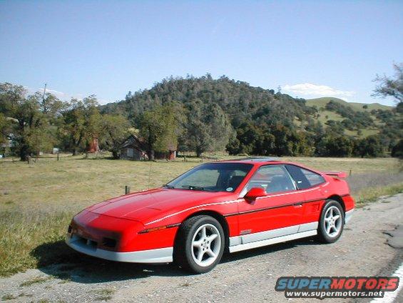 Red Pontiac Fiero parked on a dirt road with a rural landscape behind it