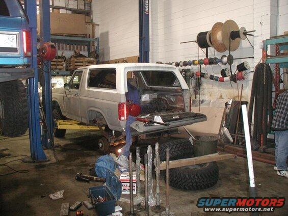 Gray Ford Bronco on a lift in a garage, tailgate open, with tools and wire spools visible.