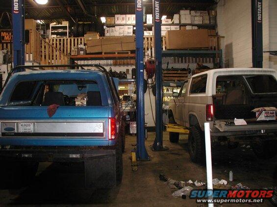 Rear view of a blue Ford Bronco next to a tan Ford Bronco on a car lift in a garage.