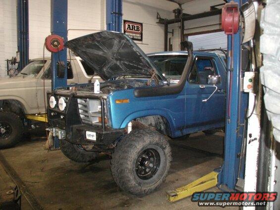 Blue 1985 Ford Bronco on a lift in a garage, hood open, with snorkel and custom bumper.