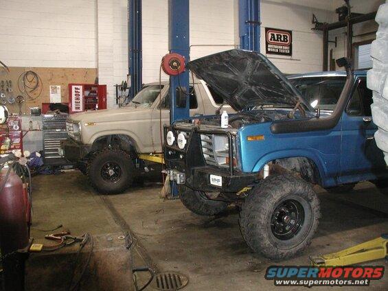 Two lifted Ford Broncos in a garage, one blue with hood open and snorkel, one tan.