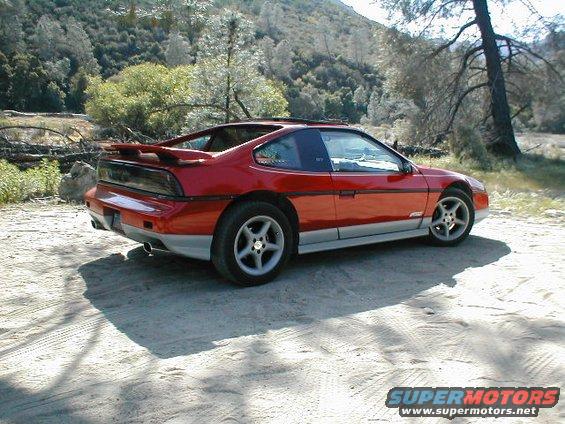 1986 Pontiac Fiero, red with silver lower trim, parked on a dirt road with trees and hills in background