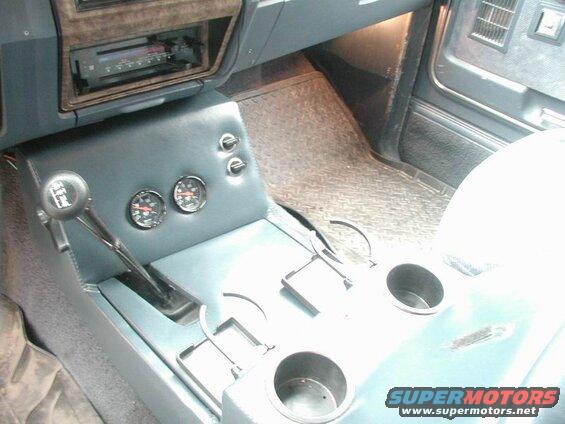 Interior of a vehicle showing a shifter, gauges, cup holders, and a car stereo.