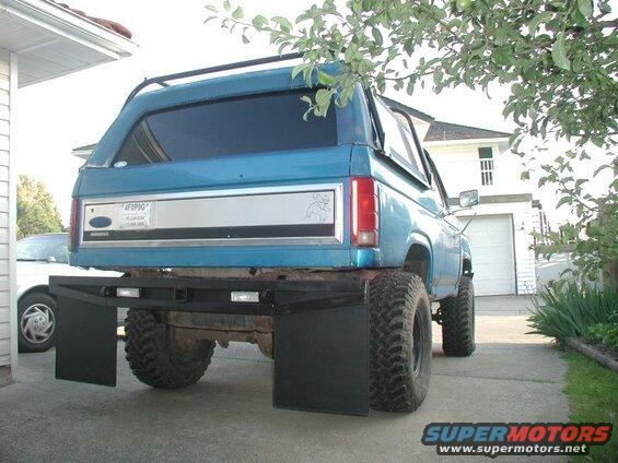 Rear view of a lifted blue 1985 Ford Bronco with large mud tires and black mud flaps.