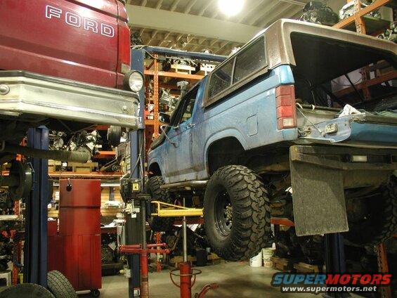 Blue Ford Bronco on a car lift in a garage with parts shelves in the background.