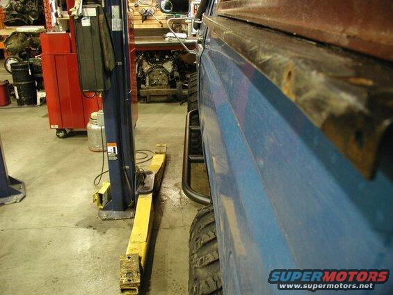 Side view of a blue vehicle's lower body and tire next to a yellow car lift in a garage.