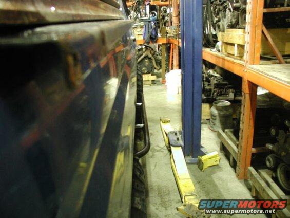 Side view of a dark blue vehicle fender and tire in a workshop with a hydraulic jack and engine parts.