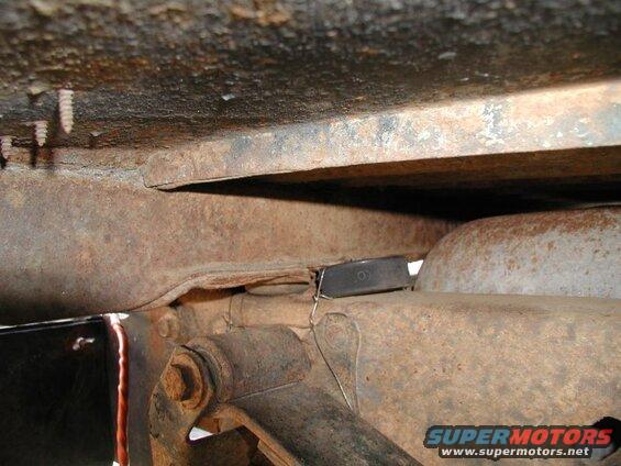 Underside of rusty vehicle frame with a black object wired to a bracket.