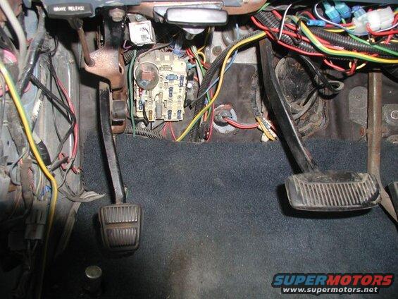 Interior view of vehicle footwell with pedals, fuse box, and wiring.