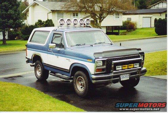 Blue and white 1985 Ford Bronco with Hella light bar and bull bar parked on wet driveway.