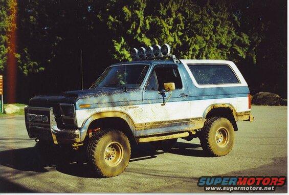 1985 Ford Bronco, blue and white, muddy, off-road tires, light bar, parked in front of trees.