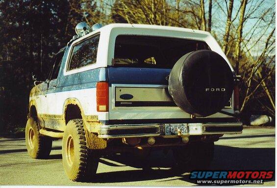 Rear view of a lifted, muddy blue and white Ford Bronco with a spare tire on the back.