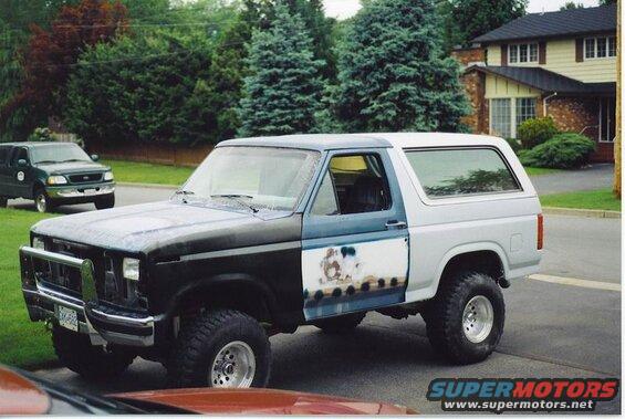 1985 Ford Bronco with primer on the driver's door, parked on a street.