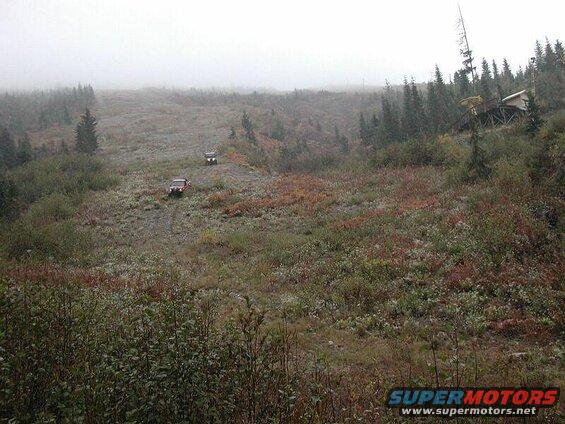 Two off-road vehicles on a muddy, rocky hill with a wooden structure on the right.