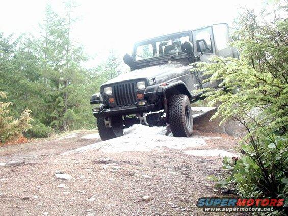 Dark Jeep Wrangler off-roading on a rocky trail with trees.