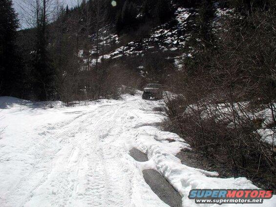 1985 Ford Bronco driving on a snowy dirt road in a wooded mountain area.