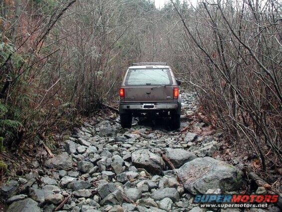 Brown Ford Bronco on a rocky, wooded trail, viewed from the rear.