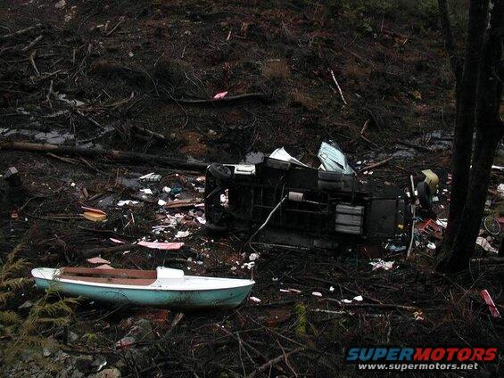 Small light blue and white boat on the ground with a wrecked vehicle and debris in the background.