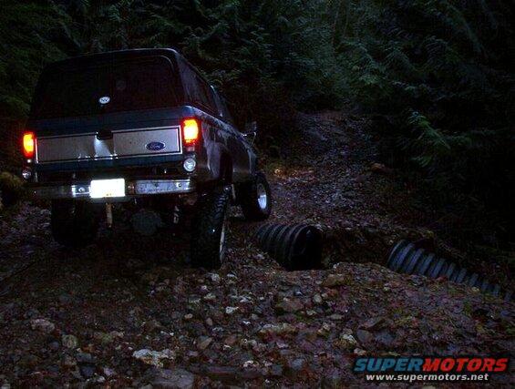 Rear view of a dark blue Ford Bronco on a muddy trail at night, brake lights on.