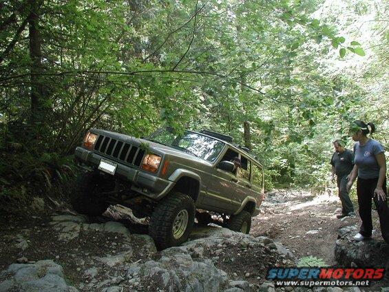 Jeep Cherokee XJ off-roading on a rocky trail in a forest.