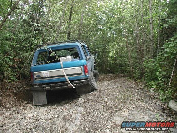 Blue 1985 Ford Bronco on a rocky, wooded trail, rear view.